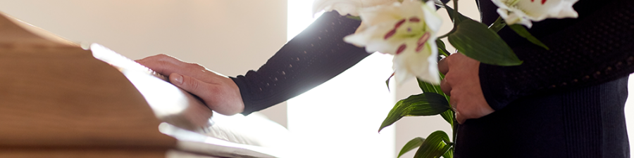 Leaseholder dies: What happens to a leasehold property when you die? A guide on extending your lease from SAM Conveyancing. A person dressed in black holds a bunch of white lilies, while placing their hand on a wooden casket.